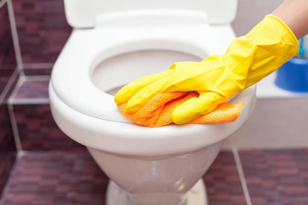 Hand in yellow glove cleaning toilet seat with orange cloth.